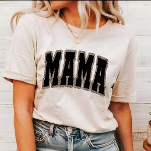 Może przedstawiać: Biała koszulka z nadrukiem słowa "MAMA" w kolorze czarnym z konturem w stylu lamparta.