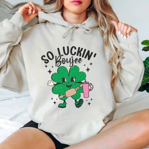 May include: A light gray hoodie with a green four-leaf clover cartoon character wearing a brown fanny pack and holding a pink tumbler. The text "SO LUCKIN' Boujee" is printed above the clover.