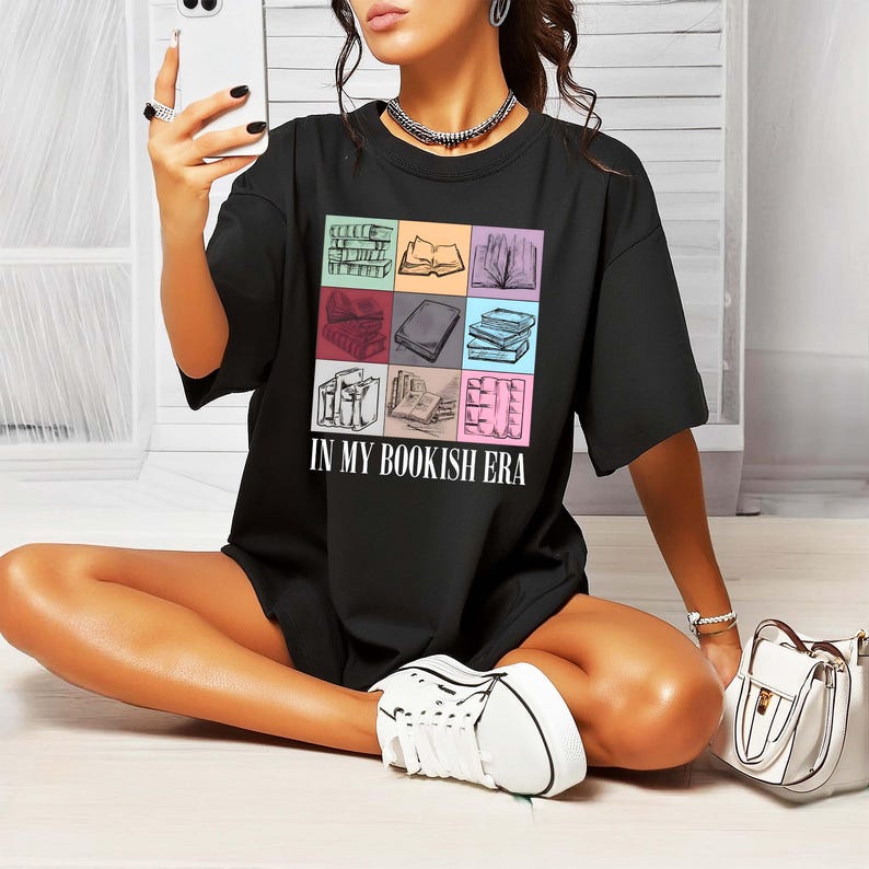 K&ouml;nnte beinhalten: Schwarzes T-Shirt mit einem farbenfrohen quadratischen Grafikdesign von B&uuml;chern und dem Text "IN MY BOOKISH ERA". Das Shirt hat einen l&auml;ssigen, &uuml;bergro&szlig;en Stil.