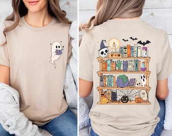 May include: Beige t-shirt featuring a cartoon ghost reading a book. The back of the shirt displays a bookshelf with Halloween-themed items like a skull, potion bottles, and a jack-o'-lantern. The design is colorful and whimsical.