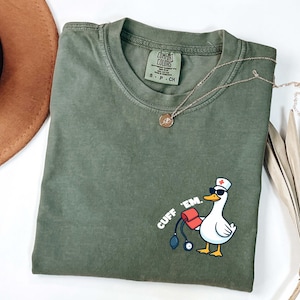 K&ouml;nnte beinhalten: Olivgr&uuml;nes T-Shirt mit einer Cartoon-Ente, die eine Sonnenbrille und eine Krankenschwesterm&uuml;tze tr&auml;gt und eine Blutdruckmanschette und ein Stethoskop h&auml;lt. Der Text "CUFF 'EM." ist auf das Shirt gedruckt. Eine goldene Halskette liegt &uuml;ber dem Shirt.