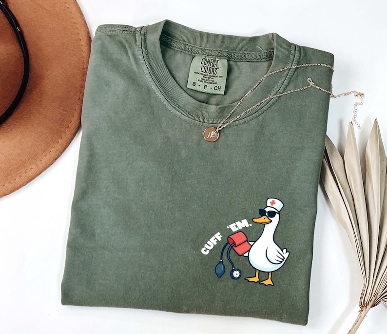 K&ouml;nnte beinhalten: Olivgr&uuml;nes T-Shirt mit einer Cartoon-Ente, die eine Sonnenbrille und eine Krankenschwesterm&uuml;tze tr&auml;gt und eine Blutdruckmanschette und ein Stethoskop h&auml;lt. Der Text "CUFF 'EM." ist auf das Shirt gedruckt. Eine goldene Halskette liegt &uuml;ber dem Shirt.