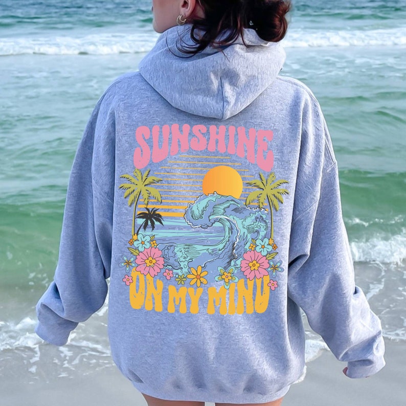 Puede incluir: Una sudadera con capucha gris claro con un gr&aacute;fico colorido en la espalda. El gr&aacute;fico presenta una escena de puesta de sol con una ola, palmeras y las palabras "SUNSHINE ON MY MIND" en rosa y amarillo. La sudadera tiene capucha y mangas largas.