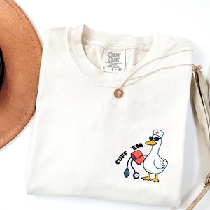 K&ouml;nnte beinhalten: Cremefarbenes T-Shirt mit einer Cartoon-Ente, die eine Sonnenbrille und eine Krankenschwesterm&uuml;tze tr&auml;gt und ein Blutdruckmessger&auml;t h&auml;lt. Der Text "CUFF 'EM." ist auf dem Shirt aufgedruckt. Eine goldene Halskette und ein brauner Hut sind ebenfalls zu sehen.