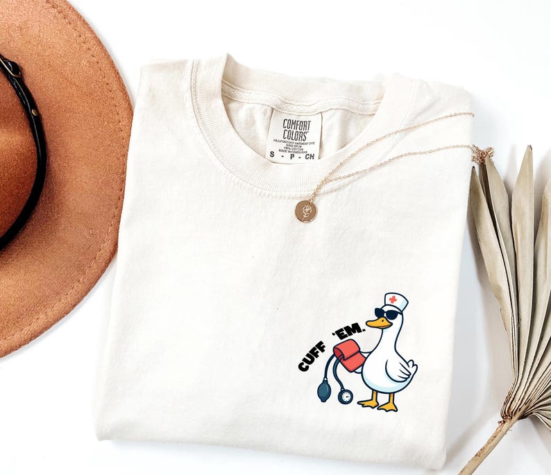 K&ouml;nnte beinhalten: Cremefarbenes T-Shirt mit einer Cartoon-Ente, die eine Sonnenbrille und eine Krankenschwesterm&uuml;tze tr&auml;gt und ein Blutdruckmessger&auml;t h&auml;lt. Der Text "CUFF 'EM." ist auf dem Shirt aufgedruckt. Eine goldene Halskette und ein brauner Hut sind ebenfalls zu sehen.