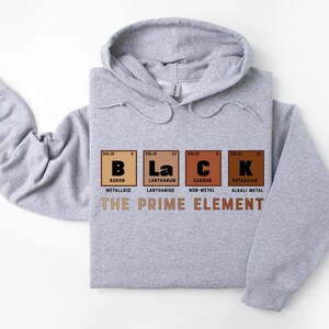 Puede incluir: Una sudadera con capucha gris con la palabra "BLACK" escrita usando los s&iacute;mbolos qu&iacute;micos del boro, lantano, carbono y potasio. El texto "THE PRIME ELEMENT" est&aacute; impreso debajo de los s&iacute;mbolos qu&iacute;micos.