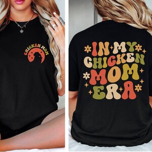 May include: Black t-shirt with a retro-style graphic that says "In My Chicken Mom Era" in colorful letters.