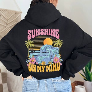 Puede incluir: Sudadera con capucha negra con un gr&aacute;fico colorido en la espalda. El dise&ntilde;o presenta un atardecer, una ola del oc&eacute;ano, palmeras y flores, con el texto "SUNSHINE ON MY MIND" en rosa y amarillo.