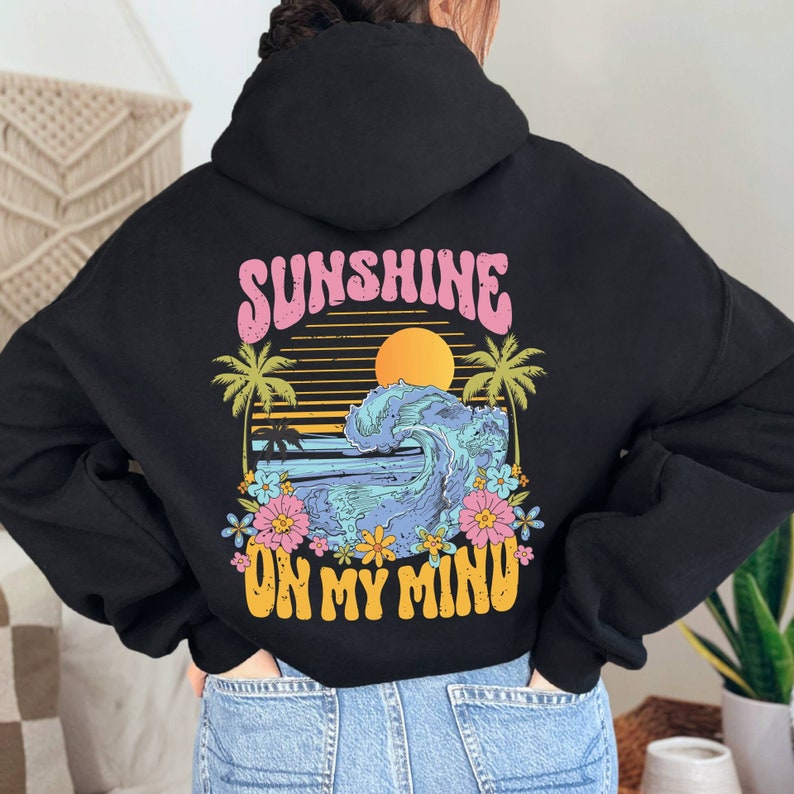 Puede incluir: Sudadera con capucha negra con un gr&aacute;fico colorido en la espalda. El dise&ntilde;o presenta un atardecer, una ola del oc&eacute;ano, palmeras y flores, con el texto "SUNSHINE ON MY MIND" en rosa y amarillo.