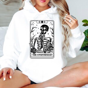May include: White hoodie with a black and white graphic of a skeleton sitting with the text "THE OVERTHINKER" on a tarot card.