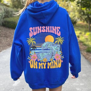 Puede incluir: Sudadera con capucha azul real con un gr&aacute;fico en la espalda. El gr&aacute;fico presenta una puesta de sol, una ola del oc&eacute;ano, palmeras y flores, con el texto "SUNSHINE ON MY MIND" en rosa y amarillo.