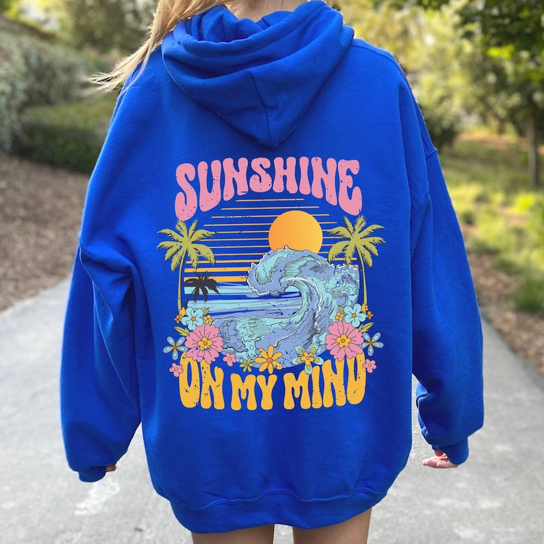 Puede incluir: Sudadera con capucha azul real con un gr&aacute;fico en la espalda. El gr&aacute;fico presenta una puesta de sol, una ola del oc&eacute;ano, palmeras y flores, con el texto "SUNSHINE ON MY MIND" en rosa y amarillo.