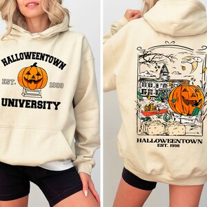 May include: Cream-colored hooded sweatshirt with "Halloweentown University" and a jack-o'-lantern graphic on the front. The back features a detailed illustration of a Halloween scene with a pumpkin and the text "Halloweentown Est. 1998".