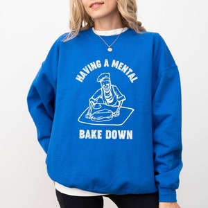 May include: A royal blue sweatshirt with a white graphic of a skeleton baking. The text on the sweatshirt reads "Having a mental bake down."