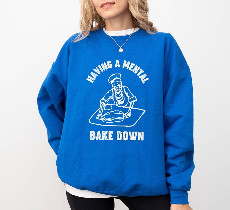 May include: A royal blue sweatshirt with a white graphic of a skeleton baking. The text on the sweatshirt reads "Having a mental bake down."