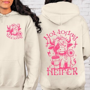 Może przedstawiać: Kremowa bluza z kapturem z r&oacute;żowym rysunkiem krowy szkockiej trzymającej kubek kawy i słonecznik. Na plecach bluzy nadrukowany jest napis "Not today Heifer".