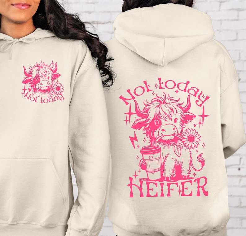 Może przedstawiać: Kremowa bluza z kapturem z r&oacute;żowym rysunkiem krowy szkockiej trzymającej kubek kawy i słonecznik. Na plecach bluzy nadrukowany jest napis "Not today Heifer".