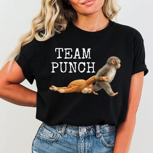 Peut inclure: T-shirt noir avec l'inscription blanche "TEAM PUNCH". Le motif représente deux singes, l'un brun et l'autre gris, dans une pose ludique. Le singe gris semble tirer le singe brun.