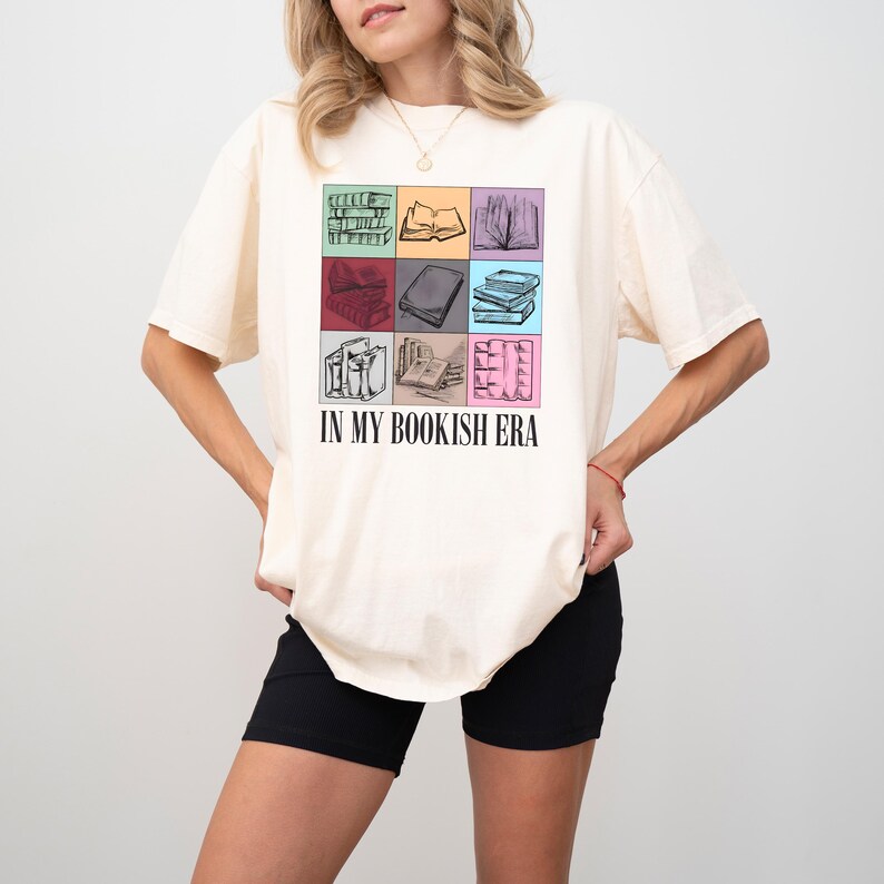 K&ouml;nnte beinhalten: Cremefarbenes T-Shirt mit einem Raster aus farbenfrohen Buchillustrationen. Das Shirt tr&auml;gt den Text "IN MY BOOKISH ERA" unter dem Raster. Das Design umfasst Bilder von offenen und geschlossenen B&uuml;chern in verschiedenen Farben.