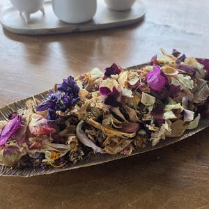 Puede incluir: Una bandeja de metal dorado con forma de hoja larga y delgada con una superficie texturizada. La bandeja está llena de una mezcla de pétalos de flores secas en tonos morado intenso, magenta vibrante, crema y bronceado, creando un arreglo floral.