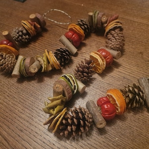 Puede incluir: Una guirnalda decorativa hecha de piñas, rodajas de naranja secas y otros elementos naturales. La guirnalda está dispuesta en forma circular y es adecuada para la decoración del hogar de temporada. Los colores incluyen marrón, naranja y verde.