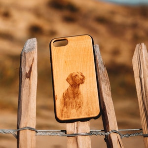 Peut inclure: Une coque de téléphone en bois avec une image gravée au laser d'un chien brun. La coque est sur une clôture en bois avec un grillage.