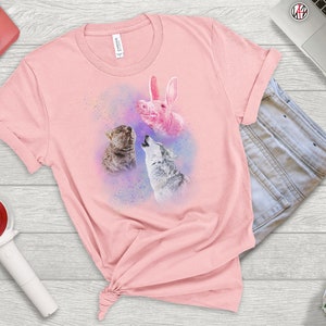 May include: Pink t-shirt with a graphic of a pink pig, a wolf, and a cat howling at the moon. The graphic is surrounded by a watercolor-like effect.