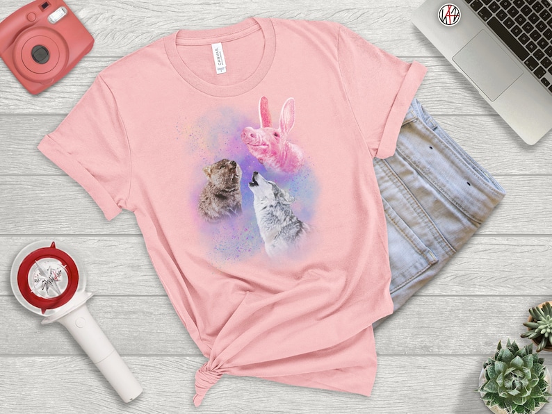 May include: Pink t-shirt with a graphic of a pink pig, a wolf, and a cat howling at the moon. The graphic is surrounded by a watercolor-like effect.