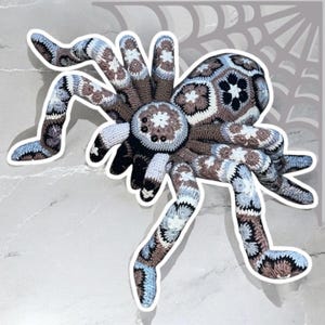 May include: A large, handmade crochet spider in shades of brown, blue, and white. The spider features a detailed pattern on its body and legs, with a circular body and eight articulated legs. The spider's eyes are small, black beads.