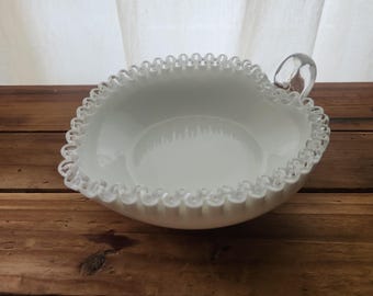 May include: A white, oval-shaped bowl with a ruffled edge and a clear glass handle. The bowl sits on a wooden surface, with a white curtain in the background. The bowl's rim has a delicate, scalloped design.