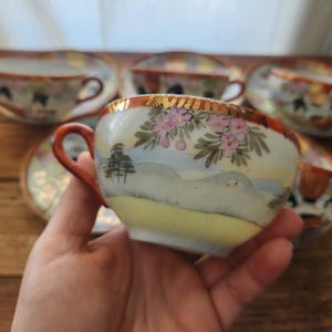 May include: A set of vintage porcelain teacups and saucers. The teacups feature a hand-painted landscape design with pink floral accents, gold trim, and a brown handle. The saucers have a matching design. The teacups are held in a person's hand.