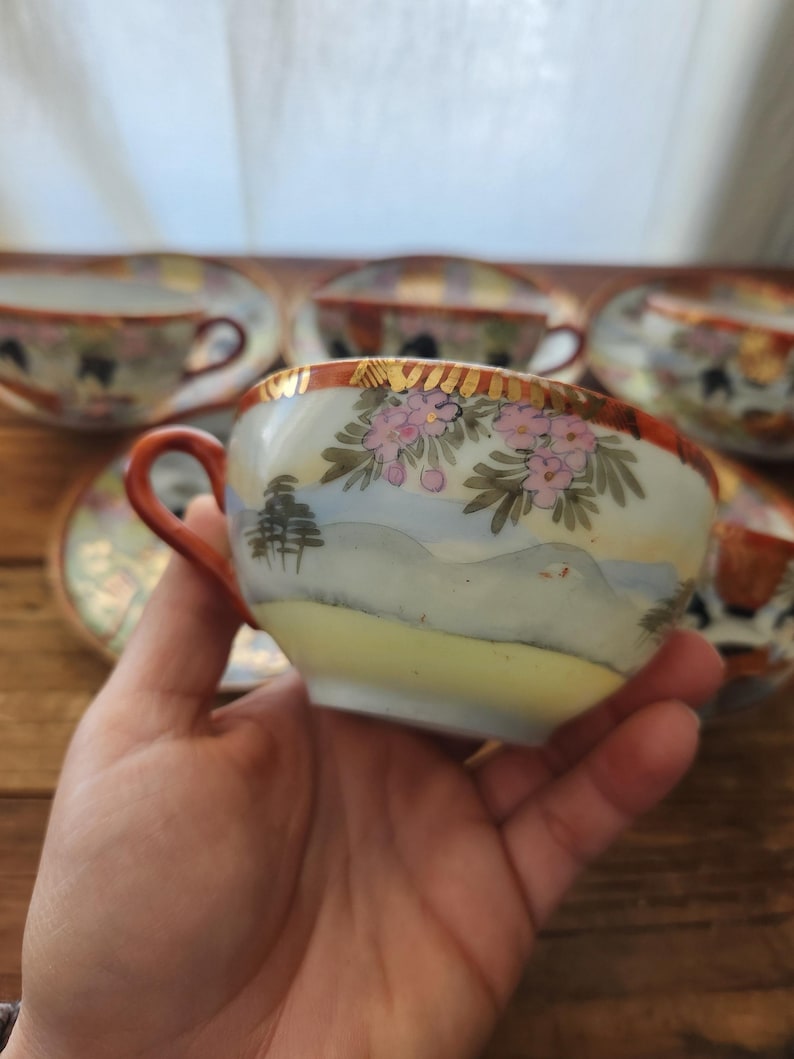 May include: A set of vintage porcelain teacups and saucers. The teacups feature a hand-painted landscape design with pink floral accents, gold trim, and a brown handle. The saucers have a matching design. The teacups are held in a person's hand.