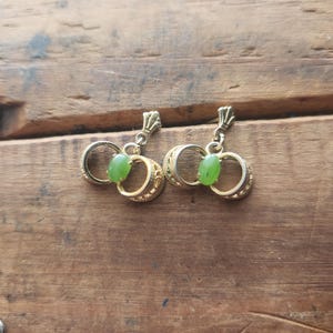 May include: A pair of gold-toned earrings with a double-hoop design and a small, oval-shaped green stone. The earrings have a decorative top and are displayed on a wooden surface. The earrings are a jewelry item.