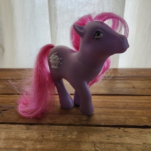 Könnte beinhalten: Lilafarbenes My Little Pony mit rosa Haaren und einem Cupcake-Symbol an der Seite.