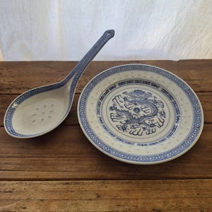 May include: A white ceramic plate and a matching slotted spoon, both decorated with blue patterns. The plate features a central blue dragon illustration surrounded by a geometric border, while the spoon handle has a matching blue design.