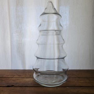 Puede incluir: Un recipiente de vidrio transparente con forma de árbol de Navidad. El recipiente tiene una parte superior puntiaguda y un diseño en capas, con una base ancha. Está sobre una superficie de madera con una cortina blanca en el fondo.