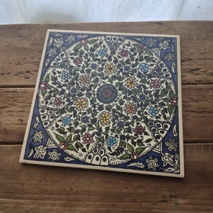 May include: A square ceramic tile featuring a central circular medallion filled with intricate floral patterns in yellow, blue, red, and green. The background is cream-colored, framed by a deep blue border with decorative white leaf motifs.