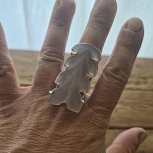 May include: A ring featuring a large, translucent, pale lavender-grey carved stone in the shape of an oak leaf. The stone is secured by four small silver-toned metal prongs that hold the leaf-shaped piece onto a simple, thin metal band.
