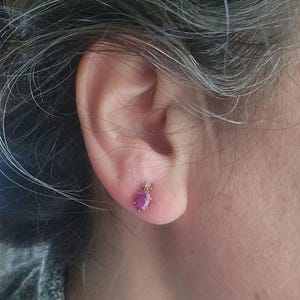 May include: A close-up of a gold stud earring with a teardrop-shaped, deep pink gemstone. A small, clear stone is set above the pink gem. The earring is set on a person's ear.