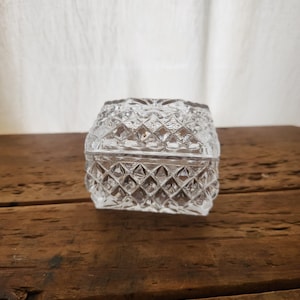 Op de afbeelding: Een heldere, vierkante glazen sieradendoos met een ruitpatroon. De doos heeft een deksel en staat op een houten oppervlak. De achtergrond is een witte stof.