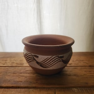 May include: A small, round terracotta pot with a wide opening. The pot features a band of white and carved wave-like designs around the middle. The pot sits on a wooden surface, with a soft, white curtain in the background. Ideal for small plants.