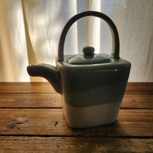 May include: A light blue and white ceramic teapot with a rounded handle. The teapot has a square body and a lid with a knob on top.