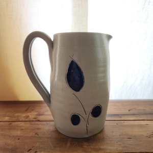 May include: A handmade ceramic pitcher with a light beige glaze and a dark blue floral design. The pitcher has a large handle and a spout for easy pouring. The design features a large leaf and two smaller circles. The pitcher is on a wooden surface.
