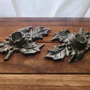 May include: Two antique metal candle holders shaped like leaves with berries. The candle holders are a dark gray color and sit on a wooden surface. Each holder has a circular opening for a candle.