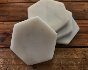 May include: Set of four white marble hexagon coasters. The coasters are stacked on top of each other on a wooden surface.