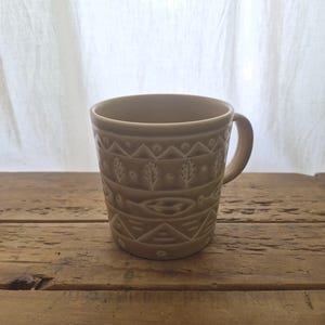 May include: A light brown ceramic mug featuring a repeating embossed pattern of horizontal bands containing triangles, small circles, and stylized leaf motifs. The mug has a smooth finish, a rounded handle on the right side, and a wide circular opening.