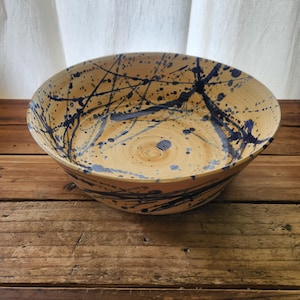 May include: A large, handmade ceramic bowl with a beige interior and exterior. The bowl is decorated with dark blue splatter paint and lines. The bowl sits on a rustic wooden surface.