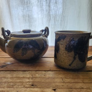 May include: A ceramic teapot and mug set. The teapot has a rounded body, a spout, and a handle. The mug has a handle. Both pieces are tan with dark blue floral designs. The set is on a wooden surface.