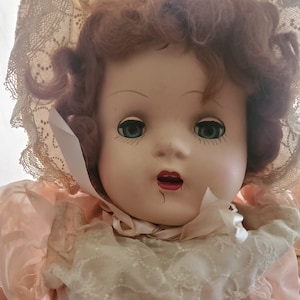 May include: A vintage porcelain doll with rosy cheeks, blue eyes, and red lips. She is wearing a pink dress with lace trim and a white lace bonnet.