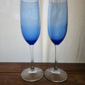 May include: Two blue glass champagne flutes with a gradient from light blue to dark blue. The flutes are on a wooden surface.